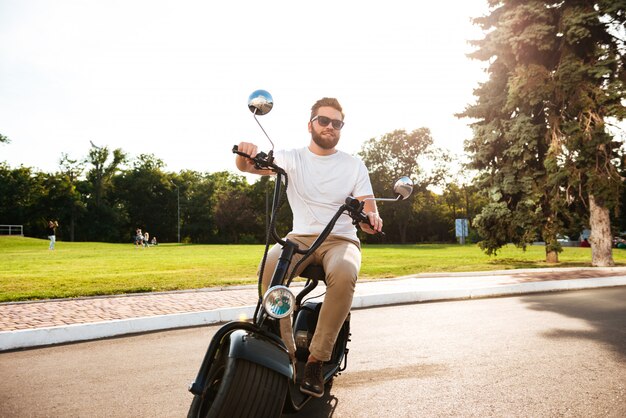 Feliz hombre con barba en gafas de sol paseos en moto moderna al aire libre y mirando a otro lado