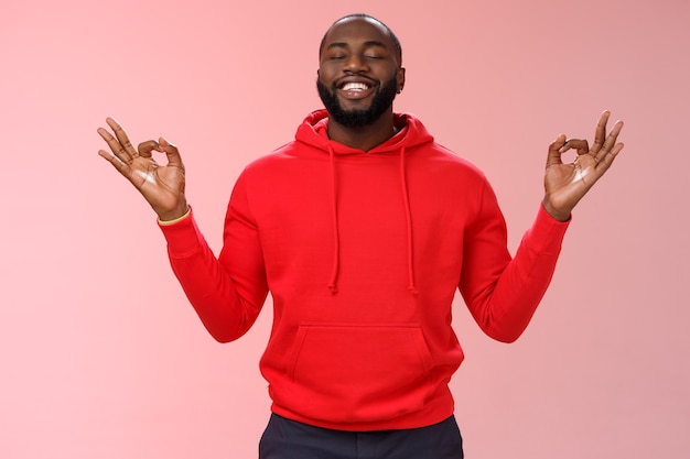 Feliz hombre barbudo afroamericano en sudadera con capucha roja meditando encontró paz nirvana sonriendo encantados ojos cerrados relajados aliviados loto de pie mudra pose buscar zen, practicar yoga, fondo rosa.