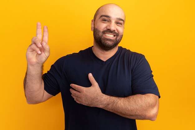 Feliz hombre barbudo en camiseta azul marino sosteniendo la mano en el pecho mostrando los dedos haciendo una promesa de pie sobre la pared naranja