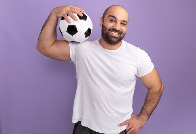 Feliz hombre barbudo en camiseta blanca sosteniendo un balón de fútbol en su hombro sonriendo de pie sobre la pared púrpura