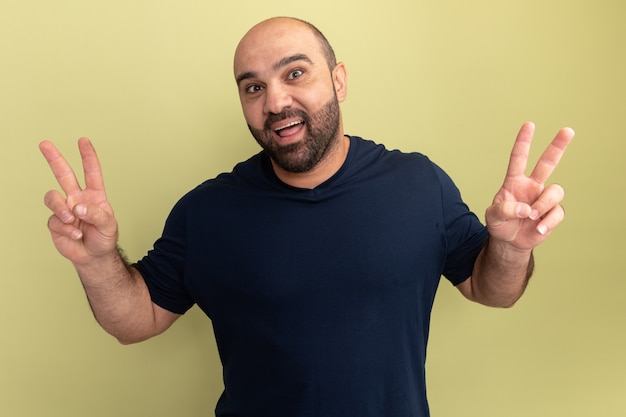 Feliz hombre barbudo en camiseta negra sonriendo alegremente mostrando v-sign de pie sobre la pared verde