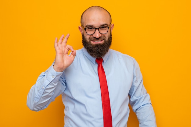 Feliz el hombre barbudo con corbata roja y camisa azul con gafas mirando con una sonrisa mostrando el signo de ok