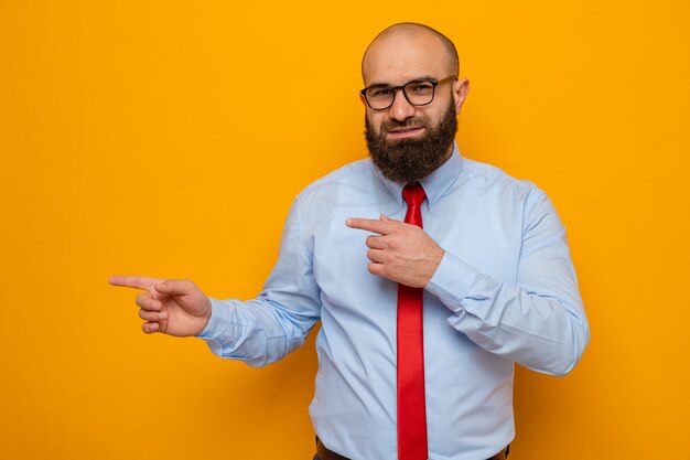 Feliz hombre barbudo con corbata roja y camisa con gafas mirando a la cámara sonriendo confiado apuntando con los dedos índices al lado de pie sobre fondo naranja