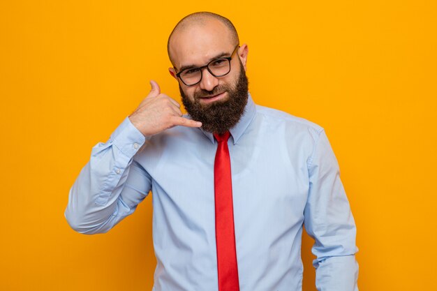 Feliz hombre barbudo con corbata roja y camisa con gafas mirando a la cámara sonriendo haciendo gesto de llamarme de pie sobre fondo naranja