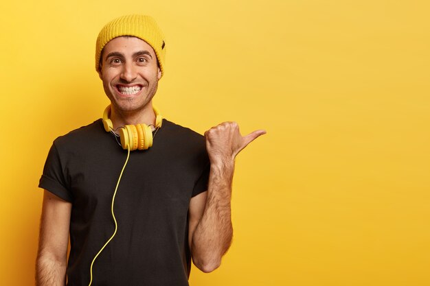 Feliz hombre caucásico positivo con sonrisa con dientes, señala con el pulgar en el espacio en blanco, promociona el elemento