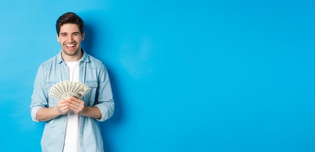 Feliz hombre exitoso sonriendo complacido sosteniendo dinero de pie sobre fondo azul.