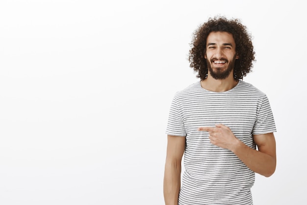 Feliz hombre guapo despreocupado con barba en elegante camiseta a rayas, riendo de alegría y apuntando hacia la izquierda con el dedo índice