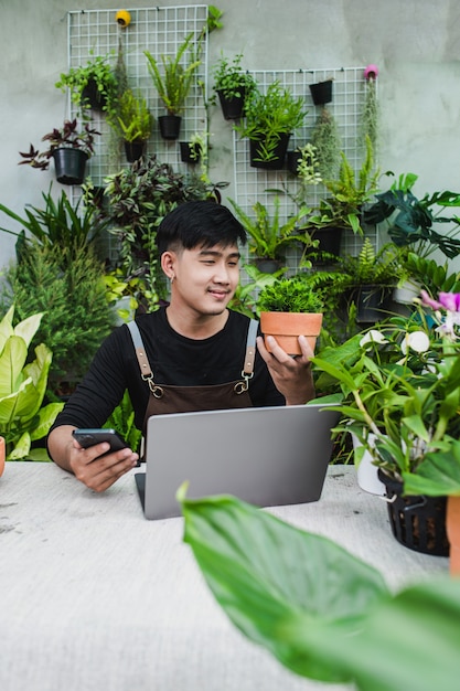 Feliz el hombre jardinero usa un teléfono inteligente y una computadora portátil mientras ofrece un tutorial en línea sobre planes en macetas en la tienda