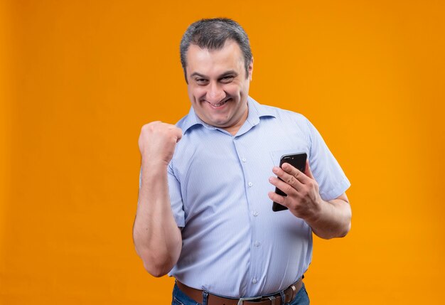 Feliz hombre de mediana edad en camisa azul de rayas verticales y levantando la mano en el gesto de puño cerrado