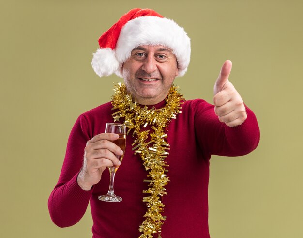 Feliz hombre de mediana edad con gorro de Papá Noel de Navidad con oropel alrededor del cuello sosteniendo una copa de champán mirando a la cámara sonriendo mostrando los pulgares para arriba sobre fondo verde