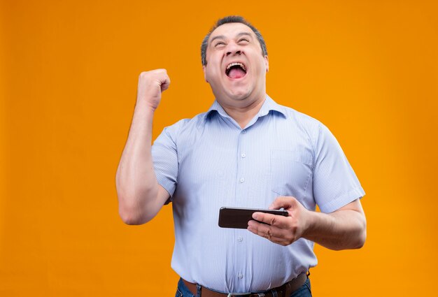 Feliz hombre de mediana edad vestido con camisa azul a rayas ganando el juego en el teléfono móvil y levantando la mano en gesto de triunfo mientras está de pie