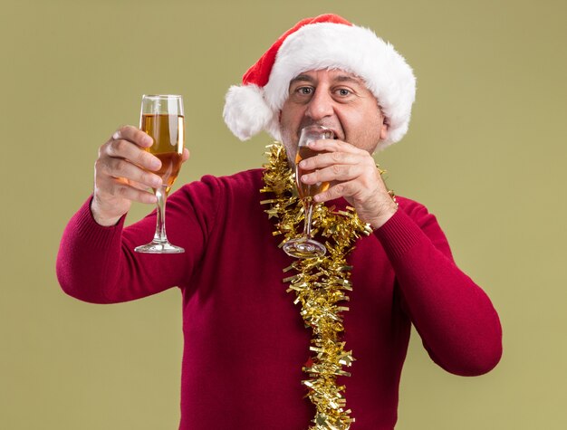 Feliz hombre de mediana edad vestido con gorro de Papá Noel de Navidad con oropel alrededor del cuello sosteniendo copas de champán bebiendo de pie sobre la pared verde