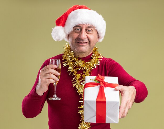 Feliz hombre de mediana edad vestido con gorro de Papá Noel de Navidad con oropel alrededor del cuello sosteniendo el regalo de Navidad y una copa de champán sonriendo alegremente de pie sobre la pared verde