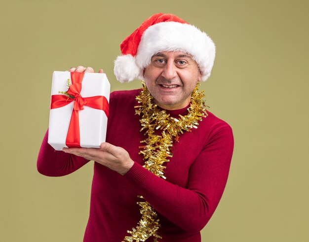 Feliz hombre de mediana edad vestido con gorro de Papá Noel de Navidad con oropel alrededor del cuello sosteniendo el regalo de Navidad mirando a la cámara con una sonrisa en la cara de pie sobre fondo verde