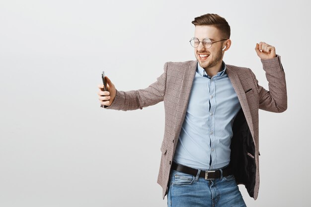 Feliz hombre de negocios bailando en regocijo con el teléfono inteligente, escuchando música en auriculares inalámbricos