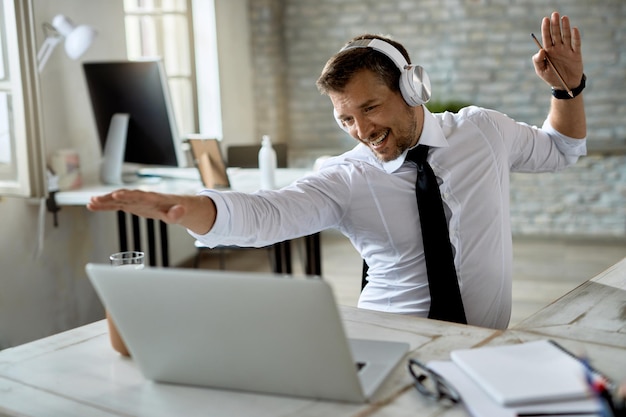 Feliz hombre de negocios escuchando música con auriculares y divirtiéndose mientras trabaja en una computadora en la oficina