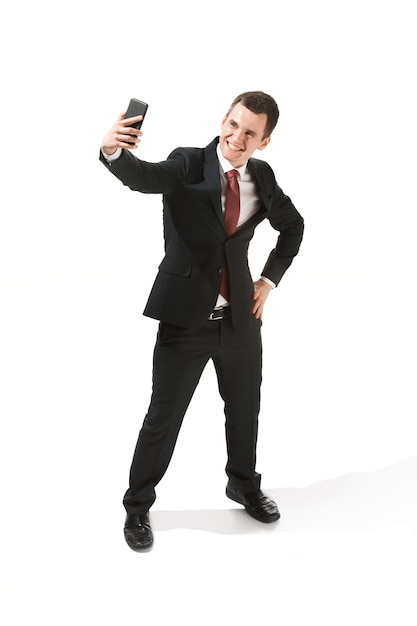 Feliz hombre de negocios hablando por teléfono sobre fondo blanco en el estudio de rodaje. Hombre joven sonriente en traje de pie y haciendo foto selfi. Negocio, carrera, concepto de éxito.