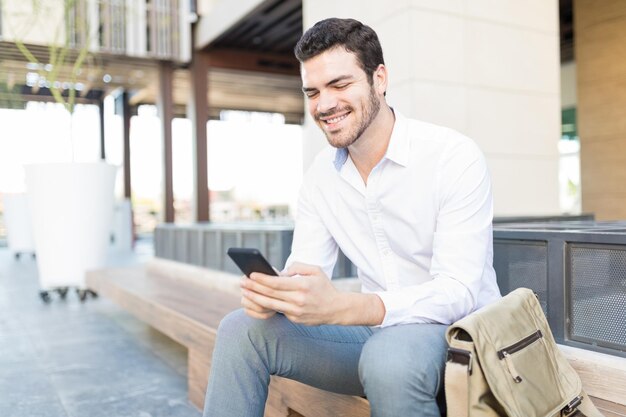 Feliz hombre de negocios hispano usando teléfono móvil mientras se sienta en un banco