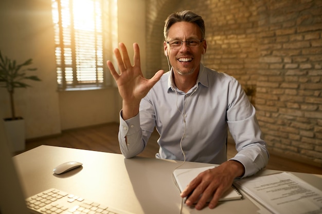 Foto gratuita feliz hombre de negocios de mediana edad saludando a alguien durante una videoconferencia en la oficina está mirando la cámara