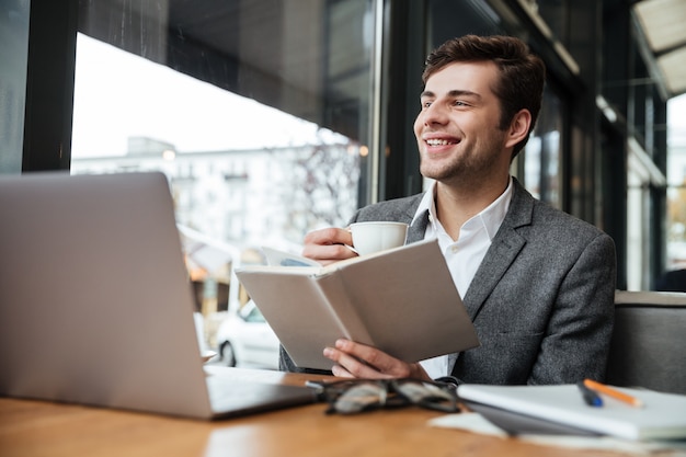 Feliz hombre de negocios sentado en la mesa de café con ordenador portátil mientras lee el libro y bebe café