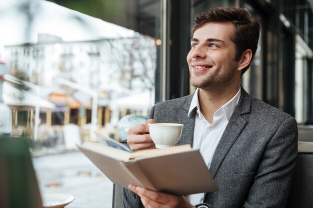 Feliz hombre de negocios sentado en la mesa de café con ordenador portátil y mirando a otro lado mientras sostiene el libro y la taza de café