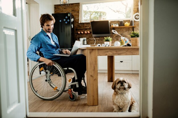 Feliz hombre de negocios en silla de ruedas mirando a su perro mientras trabaja en una laptop en casa