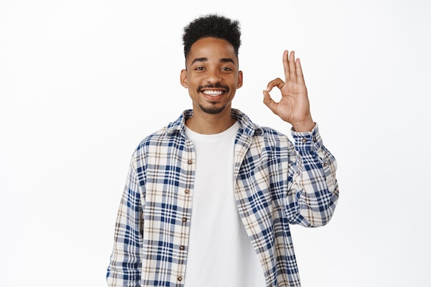 Foto gratuita feliz hombre negro sonriente de 20 años, mostrando el signo ok ok y asintiendo con aprobación, decir sí, elogiar el excelente trabajo, comentarios positivos sobre la compañía o serbio, felicitarte, antecedentes blancos