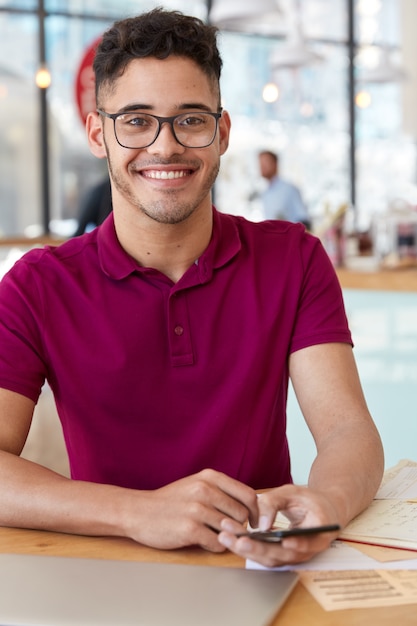 Feliz hombre de raza mixta con expresión facial alegre, charla en el teléfono móvil, conectado a internet inalámbrico, modelos contra el interior del café, tiene una sonrisa dentuda, usa camiseta casual, gafas ópticas. Blogging