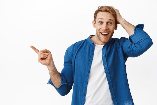 Foto gratuita feliz hombre sorprendido sonriendo, señalando con el dedo hacia la izquierda y tocando la cabeza asombrado, no puedo creer su suerte, encontró algo interesante, de pie sobre una pared blanca