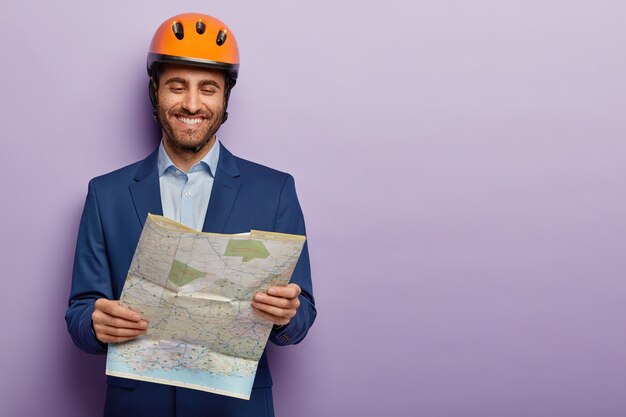 Feliz ingeniero constructor examina el plano del sitio de construcción, tiene expresión de cara alegre, usa casco, traje formal