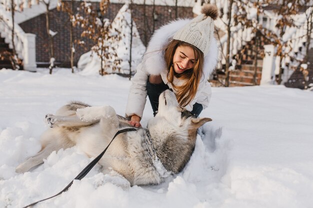 Feliz invierno de joven alegre jugando con lindo perro husky en la nieve en la calle. Estado de ánimo alegre, emociones positivas, amistad real con mascotas, amor a los animales.