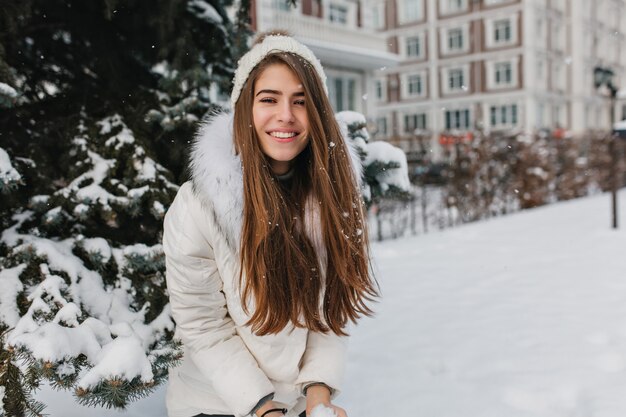Feliz invierno de mujer bonita positiva jugando con nieve. Mujer sonriente con cabello largo morena disfrutando de fin de semana durante caminar por la calle en día helado.