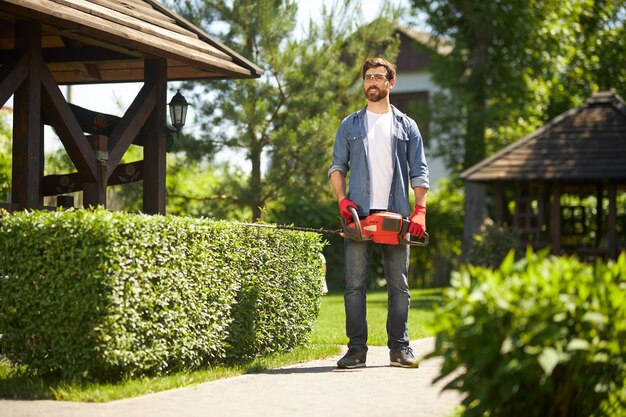 Feliz jardinero barbudo de pie llevando un moderno cortasetos de mano en el parque vista de ángulo bajo