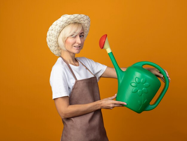 Feliz jardinero de mediana edad mujer en uniforme de jardinero con sombrero de pie en la vista de perfil sosteniendo regadera aislada en la pared naranja con espacio de copia