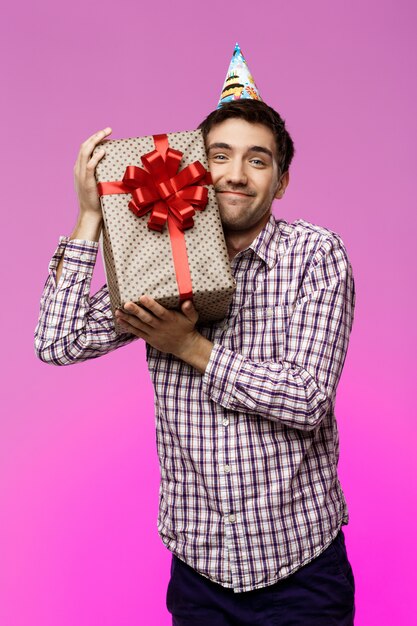 Feliz joven abrazando regalo de cumpleaños en caja sobre pared púrpura.