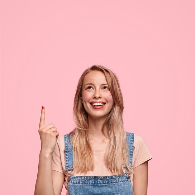 Feliz joven adolescente linda mujer con monos de mezclilla, indica hacia arriba, tiene una sonrisa positiva, se siente feliz mientras nota algo grandioso hacia arriba