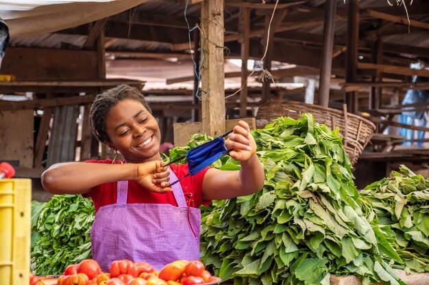 Feliz joven africana en un mercado africano local sosteniendo una mascarilla juguetonamente