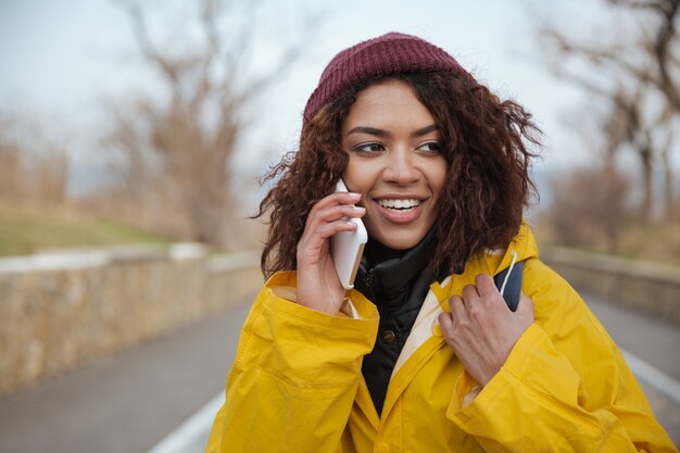 Feliz joven africana vistiendo abrigo amarillo hablando por teléfono.