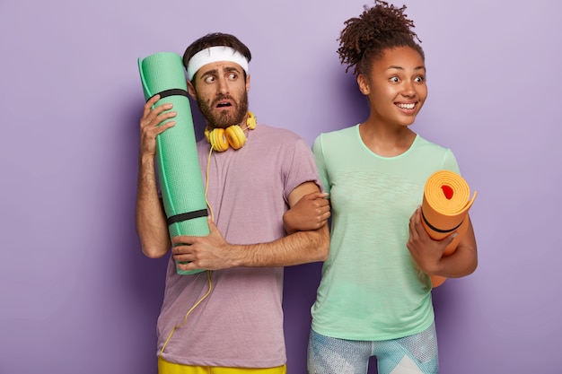 Foto gratuita feliz joven afroamericana rizada mira positivamente a un lado, toma de la mano de su novio, carga una alfombra, un hombre preocupado y desconcertado usa una diadema, usa auriculares conectados a algún dispositivo