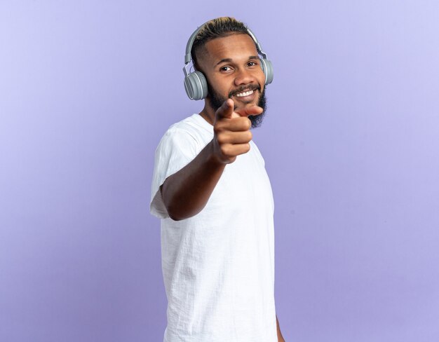 Feliz joven afroamericano en camiseta blanca con auriculares apuntando con el dedo índice a la cámara sonriendo alegremente de pie sobre fondo azul.