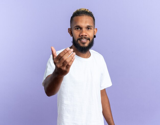 Feliz joven afroamericano en camiseta blanca mirando a la cámara sonriendo amable haciendo ven aquí gesto con la mano de pie sobre fondo azul.