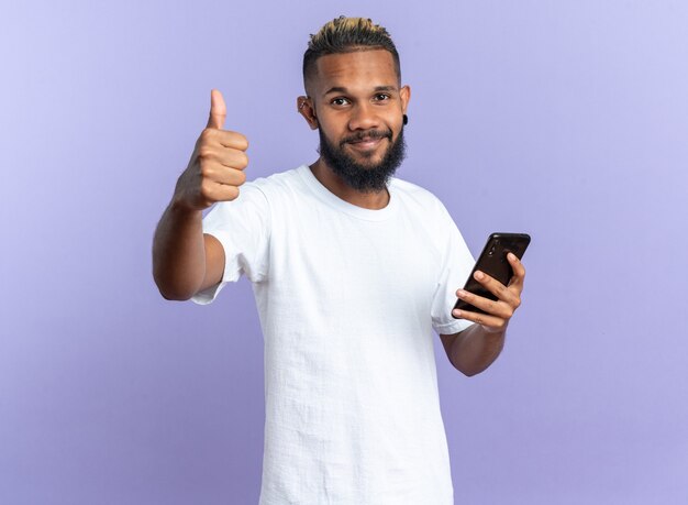 Feliz joven afroamericano en camiseta blanca sosteniendo smartphone mirando a cámara mostrando Thumbs up sonriendo alegremente