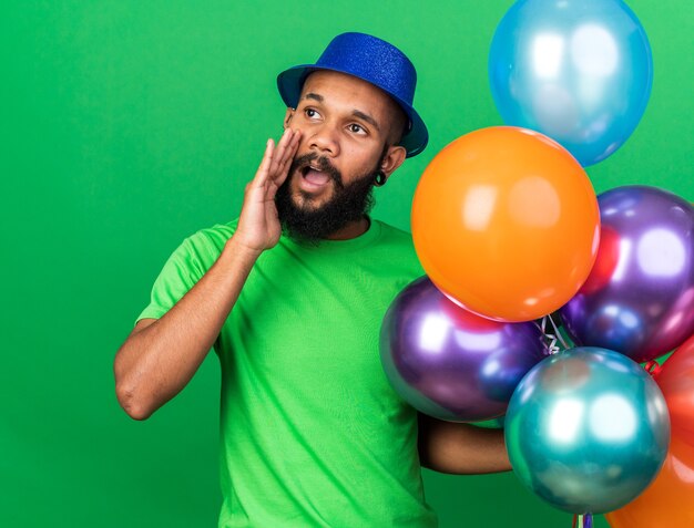 Feliz joven afroamericano con gorro de fiesta sosteniendo globos llamando a alguien