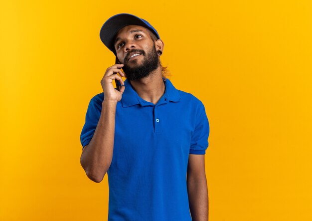 Feliz joven afroamericano repartidor hablando por teléfono mirando hacia arriba aislado sobre fondo naranja con espacio de copia