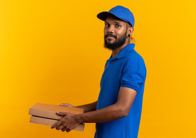 Feliz joven afroamericano repartidor de pie de lado sosteniendo cajas de pizza aisladas sobre fondo naranja con espacio de copia