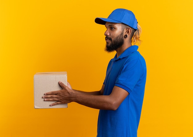 Feliz joven afroamericano repartidor de pie de lado sosteniendo el portapapeles aislado sobre fondo naranja con espacio de copia