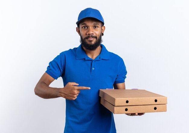 Feliz joven afroamericano repartidor sosteniendo y apuntando a cajas de pizza aisladas sobre fondo blanco con espacio de copia