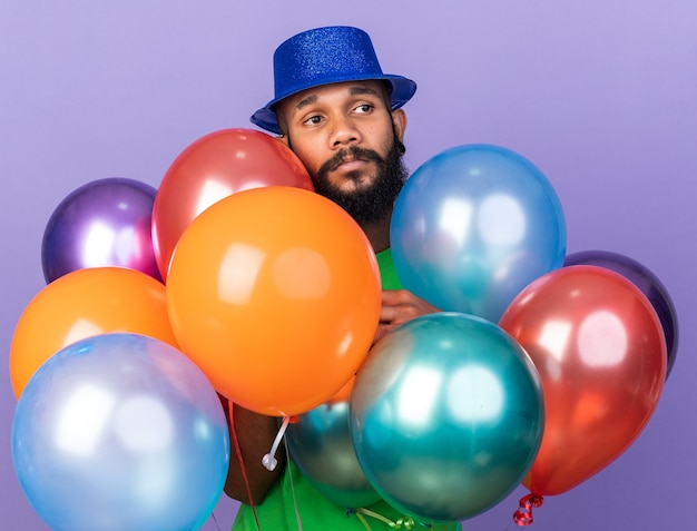 Feliz joven afroamericano con sombrero de fiesta de pie detrás de globos aislados en la pared azul