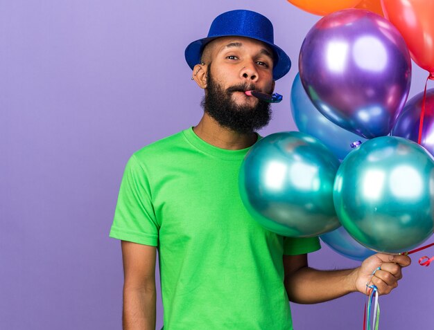 Feliz joven afroamericano vistiendo gorro de fiesta sosteniendo globos soplando silbato de fiesta aislado en la pared azul