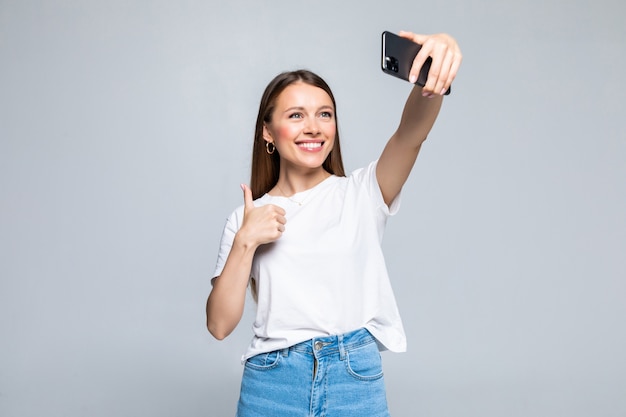 Feliz joven alegre mostrando el pulgar hacia arriba y haciendo selfie en smartphone aislado